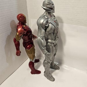 May include: Two action figures of Marvel characters. The figure on the left is Iron Man, with a red, gold, and bronze colour scheme. The figure on the right is Ultron, a silver and grey robot. Both figures are standing.