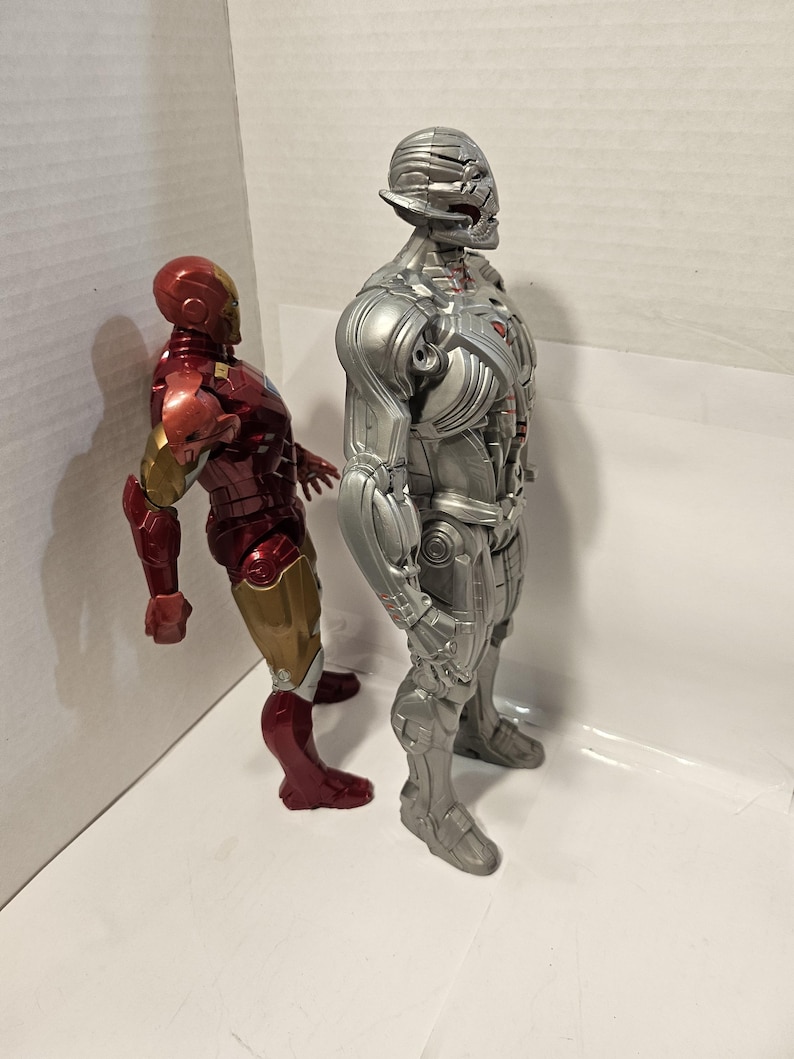 May include: Two action figures of Marvel characters. The figure on the left is Iron Man, with a red, gold, and bronze colour scheme. The figure on the right is Ultron, a silver and grey robot. Both figures are standing.