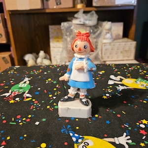 May include: A plastic figurine of Raggedy Ann, a classic children's doll, with red hair, a blue dress, and a red bow. The figurine is standing on a white base.
