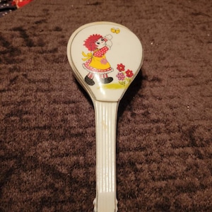 May include: A white plastic hand mirror with a handle. The mirror features a colorful illustration of a girl with red hair, wearing a yellow dress and holding a butterfly. The background is white with pink flowers.