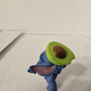 May include: A blue figurine of a cartoon character holding a green and brown avocado half. The figurine has large ears with pink insides. The avocado half is green with a brown pit.