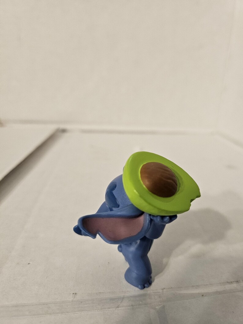 May include: A blue figurine of a cartoon character holding a green and brown avocado half. The figurine has large ears with pink insides. The avocado half is green with a brown pit.