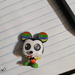 Peut inclure: Une petite figurine de Mickey Mouse noire et blanche avec des oreilles arc-en-ciel. La figurine a de grands yeux brillants et une langue rouge qui sort.