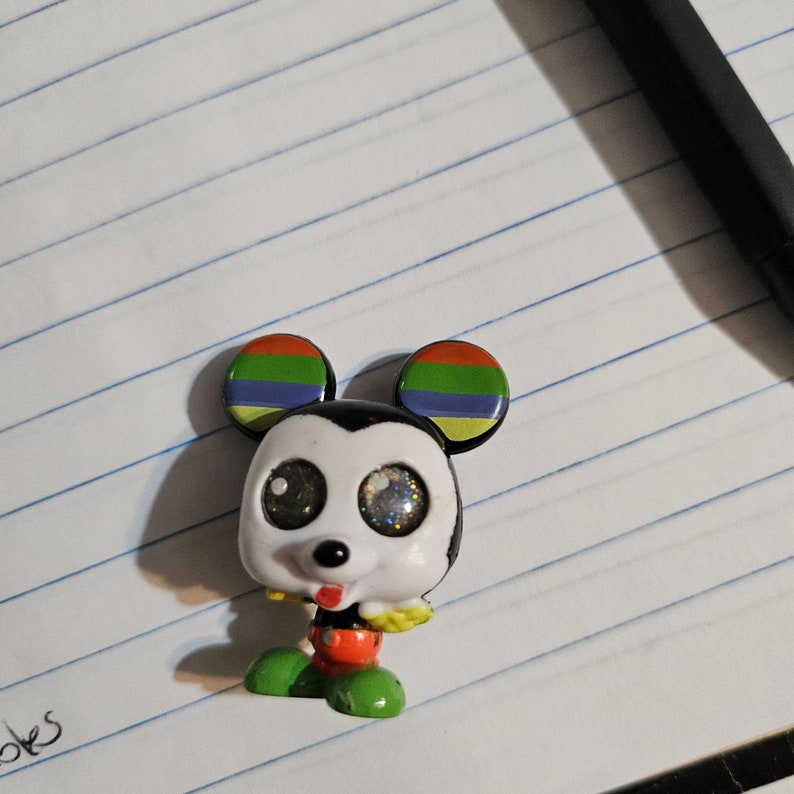 Peut inclure: Une petite figurine de Mickey Mouse noire et blanche avec des oreilles arc-en-ciel. La figurine a de grands yeux brillants et une langue rouge qui sort.