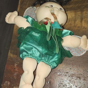 Puede incluir: Una muñeca Cabbage Patch Kid vestida con un vestido de satén verde con un diseño de hoja de acebo y detalles de cinta dorada. La muñeca tiene cabello negro y una cara pintada.