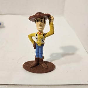 May include: A small plastic figurine of a cowboy character, wearing a brown hat, yellow shirt with black spots, a vest, blue jeans, and brown boots. The figure is standing on a brown oval base, with one hand raised to the hat.