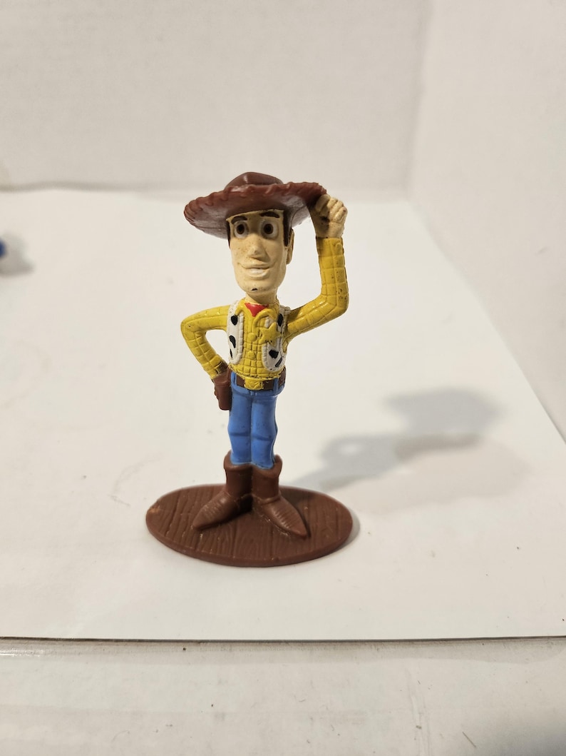 May include: A small plastic figurine of a cowboy character, wearing a brown hat, yellow shirt with black spots, a vest, blue jeans, and brown boots. The figure is standing on a brown oval base, with one hand raised to the hat.