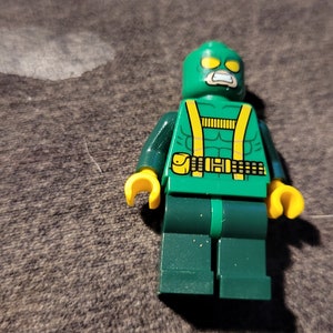 Puede incluir: Minifigura Lego verde con un cinturón amarillo y una máscara amarilla y verde. La minifigura tiene una boca blanca y ojos amarillos.