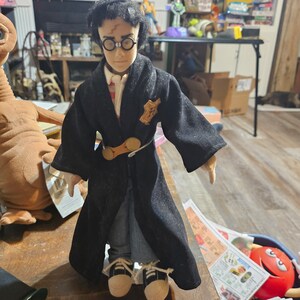 K&ouml;nnte beinhalten: Eine schwarze und braune Puppe, die eine schwarze Robe mit einem goldenen G&uuml;rtel und einem braunen und wei&szlig;en Schal tr&auml;gt. Die Puppe hat schwarze Haare und tr&auml;gt eine Brille.