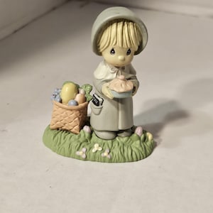 Könnte beinhalten: Figur eines Kindes in Pilgerkleidung, das einen Kuchen hält. Links befindet sich ein Korb mit Produkten. Die Figur steht auf einer grünen Basis mit Blumen. Das Kind hat blondes Haar und trägt ein graues Outfit.