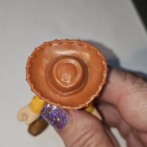 Op de afbeelding: Een bruine cowboyhoed, onderdeel van een speelgoedfiguur, wordt vastgehouden door een hand met een paarse glitter nagel. De hoed heeft een gestikte rand en een cirkelvormige inkeping aan de bovenkant. De figuur heeft een geel shirt en bruine laarzen.