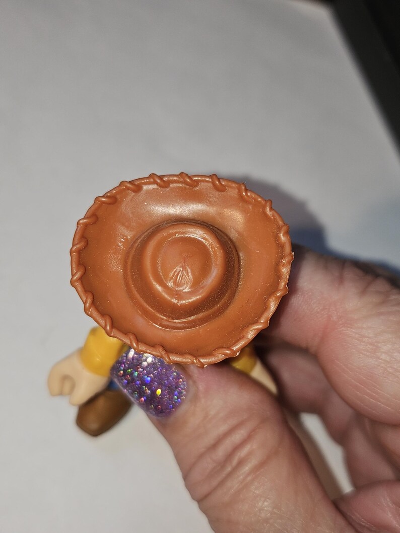 Op de afbeelding: Een bruine cowboyhoed, onderdeel van een speelgoedfiguur, wordt vastgehouden door een hand met een paarse glitter nagel. De hoed heeft een gestikte rand en een cirkelvormige inkeping aan de bovenkant. De figuur heeft een geel shirt en bruine laarzen.