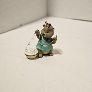 May include: A small, brown bear figurine wearing a teal dress with the word "Mom" on it. The bear holds a string of white beads and a small bowl filled with white material. A red bow adorns the bear's head.