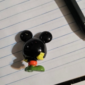 Peut inclure: Une figurine en plastique noire de Mickey Mouse avec des d&eacute;tails jaunes, rouges et verts. La figurine est debout sur une surface blanche.
