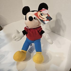May include: A plush Mickey Mouse toy with black ears, a red top, blue trousers, and yellow shoes. The toy has white gloves and a tag with Mickey's image. The tag has the text "5.99 EA".