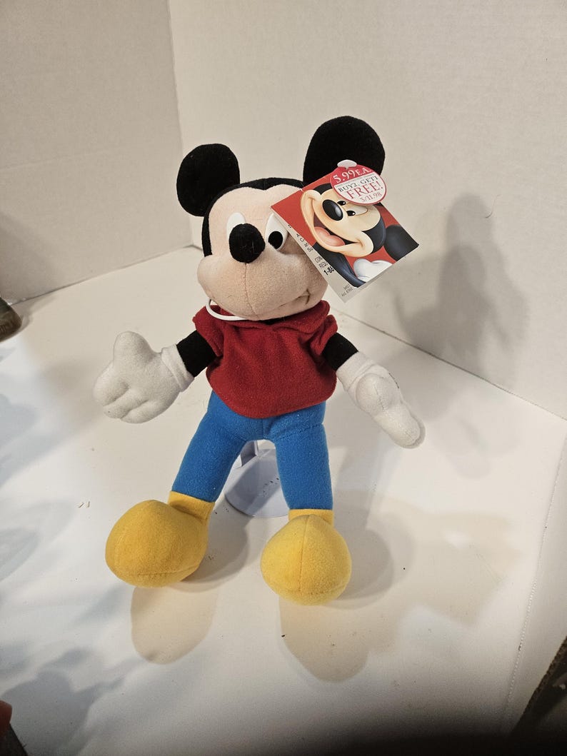 May include: A plush Mickey Mouse toy with black ears, a red top, blue trousers, and yellow shoes. The toy has white gloves and a tag with Mickey's image. The tag has the text "5.99 EA".