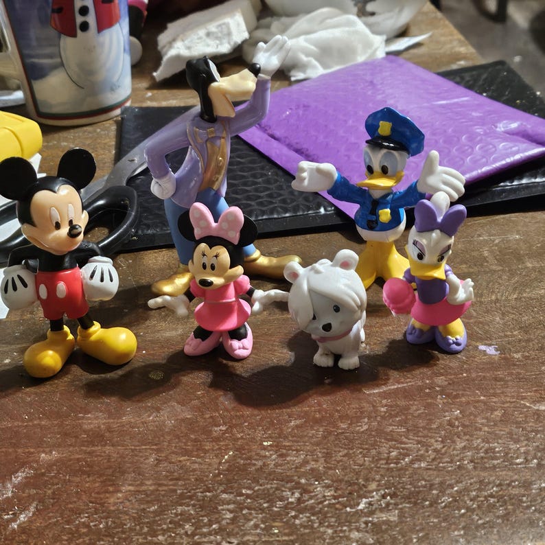 Może przedstawiać: Kolekcja figurek postaci Disneya, w tym Myszka Miki w czerwonych spodenkach, Goofy w fioletowym garniturze, Myszka Minnie w r&oacute;żowej sukience, Kaczor Donald w mundurze policjanta, Daisy i biała figurka misia. Na drewnianej powierzchni.