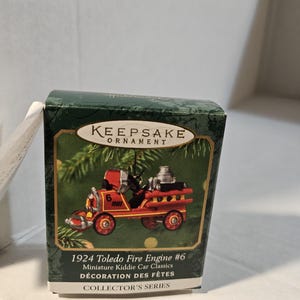Könnte beinhalten: Eine Keepsake Ornament-Box mit einer Miniatur-Feuerwehr #6 von 1924 Toledo in Rot und Silber. Die Box ist dunkelgrün mit goldfarbenen Buchstaben und einem Weihnachtsbaum-Design. Der Text "COLLECTOR'S SERIES" ist ebenfalls sichtbar.