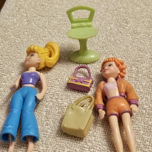 May include: Two plastic dolls, one with blonde hair wearing a purple top and blue pants, the other with orange hair wearing an orange outfit, accompanied by a light green chair, a small purple handbag, and a beige tote bag accessory.