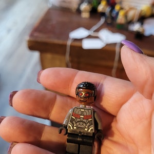 Può includere: Una minifigure Lego grigia e nera con occhiali rossi e una parrucca nera. La minifigure indossa un abito grigio e nero con accenti rossi.