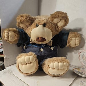 Op de afbeelding: Een pluche teddybeer met lichtbruine vacht, gekleed in een blauw geruit overhemd en een spijkerbroek. De beer heeft een grote, crèmekleurige snuit en poten. De poten van de beer zijn gesigneerd. Een decoratieve theekop is zichtbaar op de achtergrond.