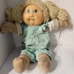 May include: A vintage Cabbage Patch Kids doll with blonde yarn hair styled in pigtails. The doll has blue eyes, rosy cheeks, and is wearing a light green corduroy jumpsuit with white lace trim.