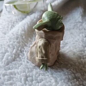 Puede incluir: Una peque&ntilde;a figura verde de un personaje de una popular franquicia de ciencia ficci&oacute;n. La figura est&aacute; en un soporte de color beige. El personaje tiene orejas grandes y mira hacia abajo. La figura est&aacute; sobre una superficie blanca texturizada.