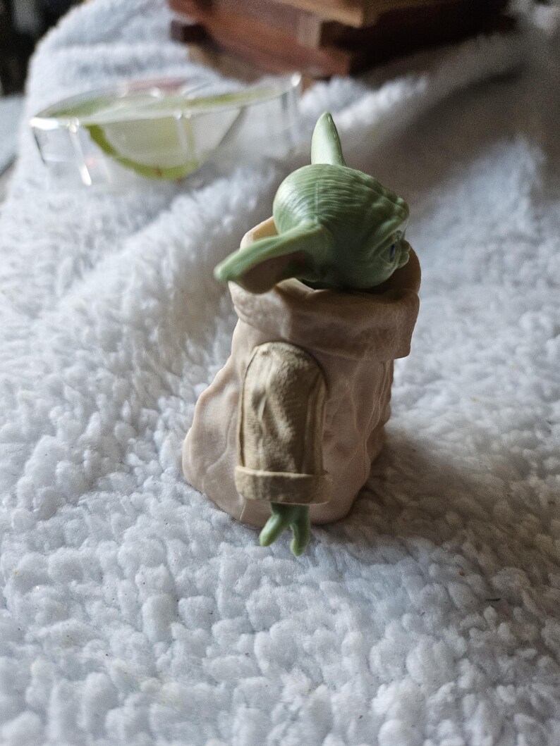 Puede incluir: Una peque&ntilde;a figura verde de un personaje de una popular franquicia de ciencia ficci&oacute;n. La figura est&aacute; en un soporte de color beige. El personaje tiene orejas grandes y mira hacia abajo. La figura est&aacute; sobre una superficie blanca texturizada.