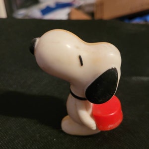 Peut inclure: Une figurine en plastique blanche de Snoopy portant un objet rond rouge. Snoopy porte un collier noir et a des yeux et des oreilles noirs.