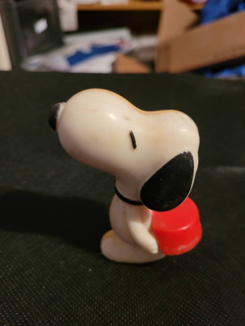 Peut inclure: Une figurine en plastique blanche de Snoopy portant un objet rond rouge. Snoopy porte un collier noir et a des yeux et des oreilles noirs.