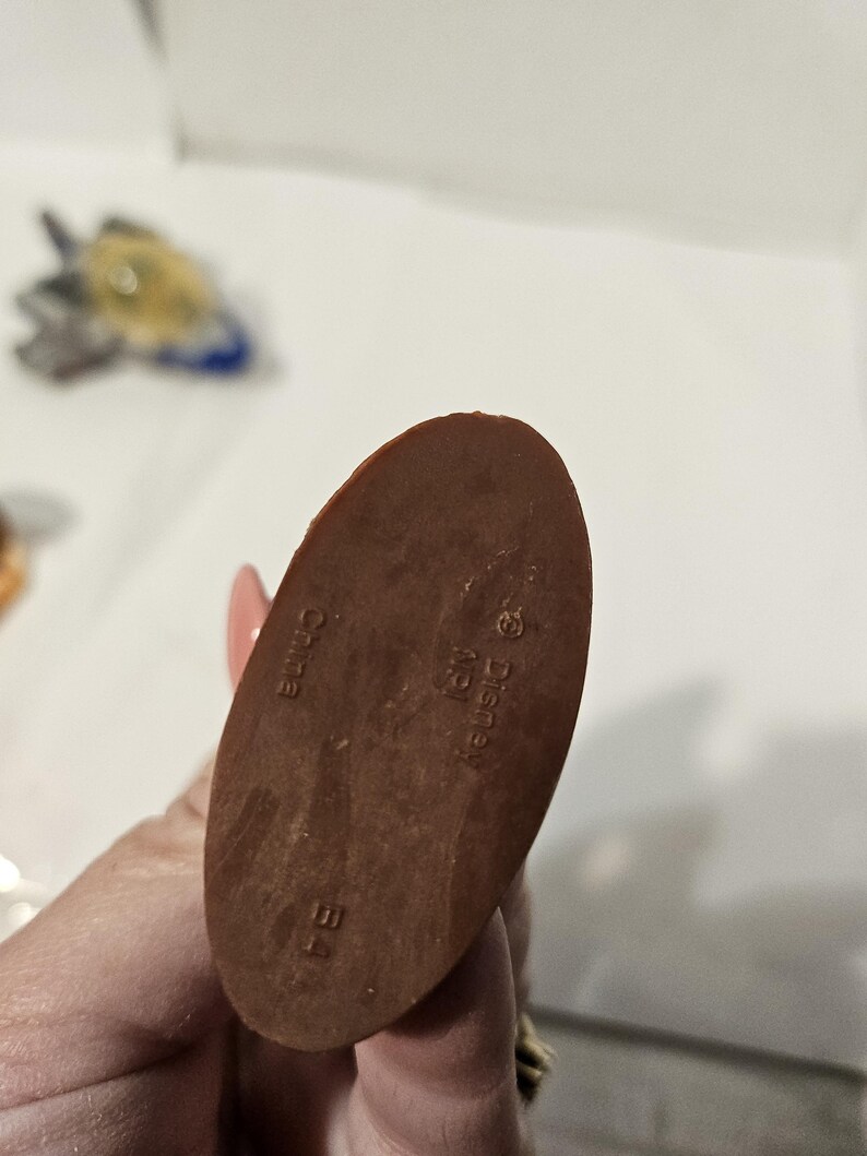 May include: A brown, oval-shaped figurine base with the text "China", "Disney", and "B4" imprinted on it. The figurine base is being held by a person.