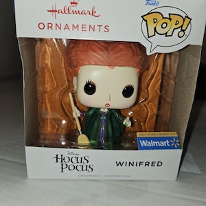 May include: A Funko Pop! vinyl figure of Winifred from Disney's Hocus Pocus, packaged in a Hallmark Ornaments box. The figure has red hair, a green dress, and holds a broom. The box features the Walmart logo.
