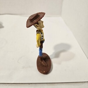 May include: A small plastic figurine of a cowboy character, wearing a brown hat, yellow shirt, blue trousers, and brown boots. The figure is standing on a brown oval base. The character is looking upwards.
