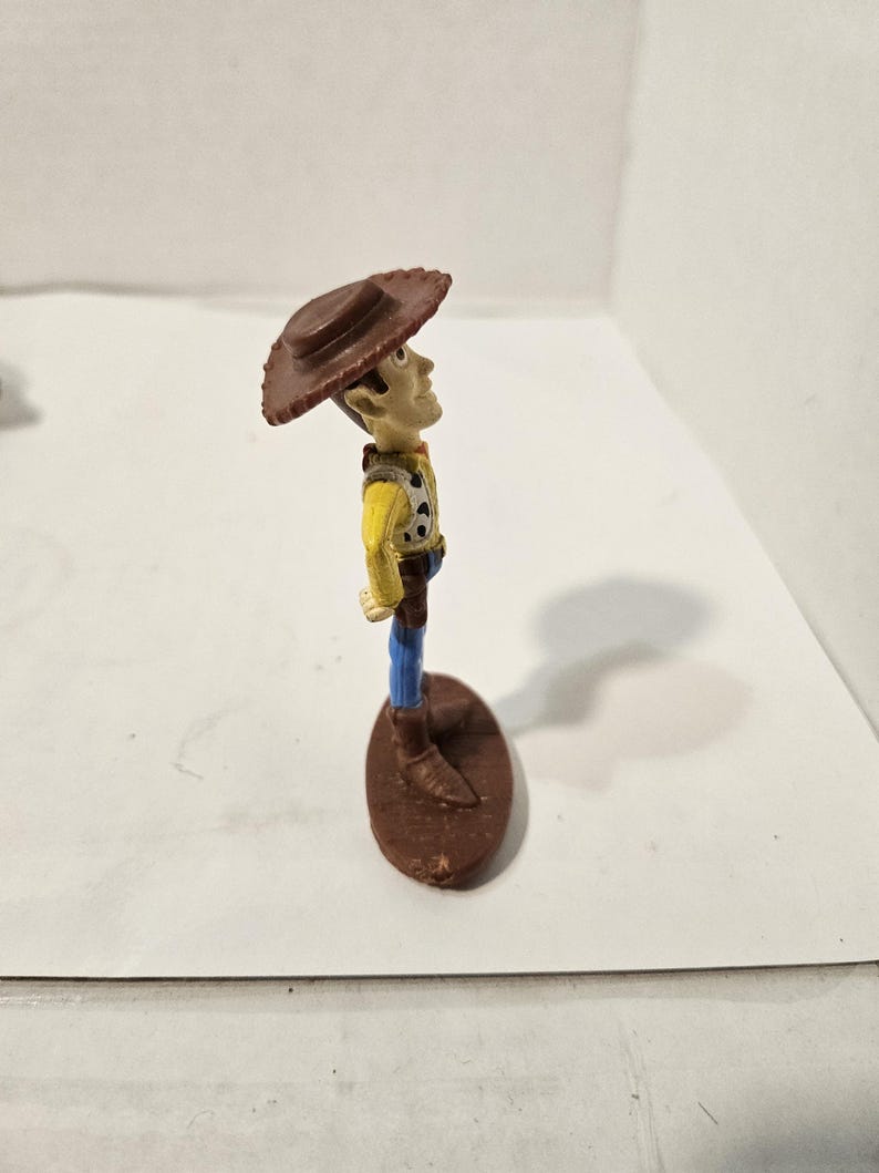 May include: A small plastic figurine of a cowboy character, wearing a brown hat, yellow shirt, blue trousers, and brown boots. The figure is standing on a brown oval base. The character is looking upwards.