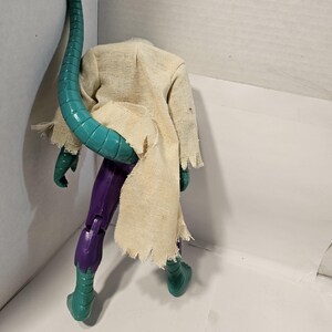 May include: A headless action figure of a lizard-like creature. The figure has a teal tail, green feet and hands, purple legs, and a tattered tan cloth draped over its shoulders. The figure is posed against a white background.
