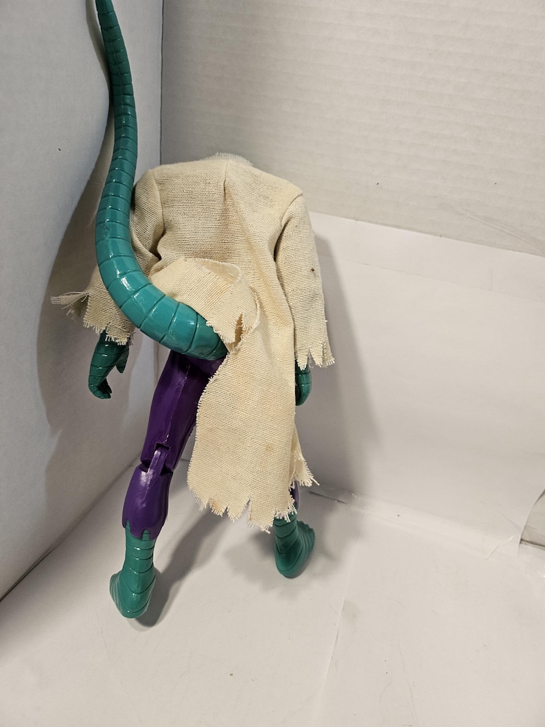 May include: A headless action figure of a lizard-like creature. The figure has a teal tail, green feet and hands, purple legs, and a tattered tan cloth draped over its shoulders. The figure is posed against a white background.