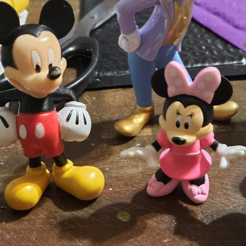 Może przedstawiać: Figurki Myszki Miki i Myszki Minnie. Myszka Miki ma na sobie czerwone spodenki, czarne rękawiczki i ż&oacute;łte buty. Myszka Minnie ma na sobie r&oacute;żową sukienkę i kokardę. Inne figurki są widoczne w tle.