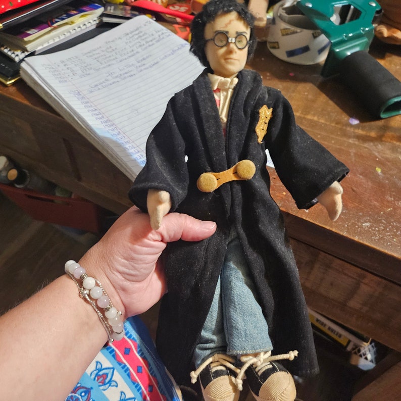 K&ouml;nnte beinhalten: Eine Pl&uuml;schpuppe von Harry Potter, die eine schwarze Robe mit einem goldenen Emblem und Jeans tr&auml;gt. Die Puppe hat schwarze Haare, braune Augen und tr&auml;gt eine Brille.