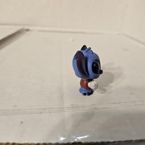 Pu&ograve; includere: Una piccola statuina blu di un personaggio dei cartoni animati con grandi occhi neri e un oggetto bianco a forma di palla. La statuina ha un top rosso e pantaloncini blu. Le orecchie del personaggio sono grandi e a punta.