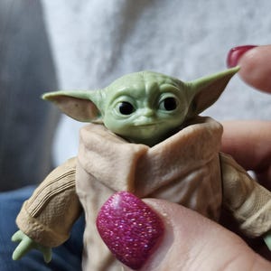 Puede incluir: Una peque&ntilde;a figura verde de un personaje con orejas grandes y ojos oscuros, que lleva un abrigo color canela. La figura se sostiene en una mano con esmalte de u&ntilde;as rosa brillante.