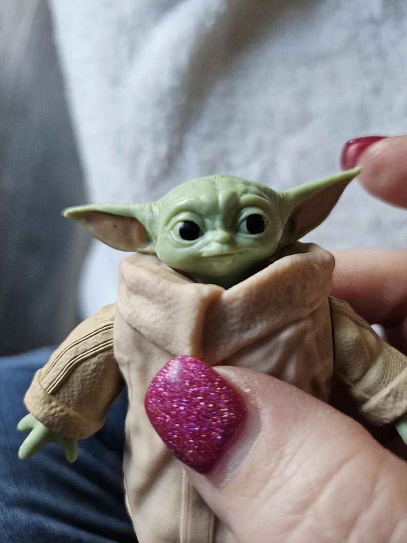Puede incluir: Una peque&ntilde;a figura verde de un personaje con orejas grandes y ojos oscuros, que lleva un abrigo color canela. La figura se sostiene en una mano con esmalte de u&ntilde;as rosa brillante.