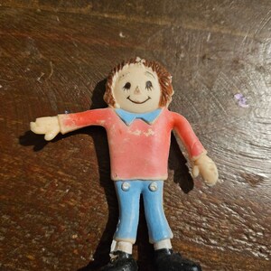 Könnte beinhalten: Eine vintage Spielfigur aus Plastik, die eine Person mit braunen Haaren darstellt, die ein rotes Hemd, eine blaue Jeans und schwarze Schuhe trägt. Die Figur hat ein lächelndes Gesicht mit bemalten Augen und einem Mund.