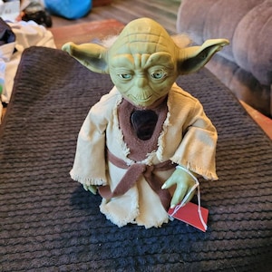 K&ouml;nnte beinhalten: Eine gr&uuml;ne Yoda-Figur, die eine braune Robe mit einem braunen G&uuml;rtel tr&auml;gt. Die Figur steht auf einer schwarzen, strukturierten Oberfl&auml;che.