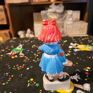 May include: A red-haired doll wearing a blue dress with white trim and red polka dots. The doll is holding a white and red object in her right hand. The doll is standing on a white base.
