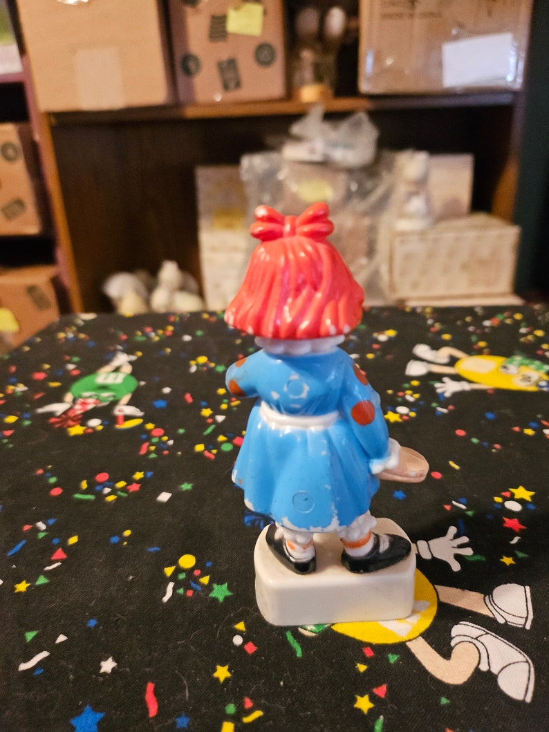 May include: A red-haired doll wearing a blue dress with white trim and red polka dots. The doll is holding a white and red object in her right hand. The doll is standing on a white base.
