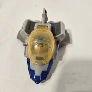 May include: A toy spaceship with a clear yellow cockpit and a figure inside. The spaceship is primarily gray with blue and white accents. The toy is likely a collectible or a children's plaything.
