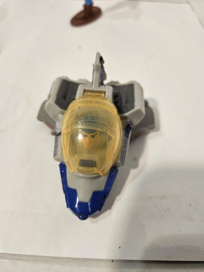 May include: A toy spaceship with a clear yellow cockpit and a figure inside. The spaceship is primarily gray with blue and white accents. The toy is likely a collectible or a children's plaything.
