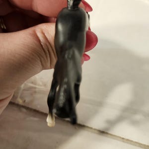 Puede incluir: Pequeña figura de caballo negro con una pezuña blanca. El caballo está en posición encabritada, con la cabeza y la cola levantadas. La figura está hecha de un material liso y brillante.