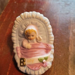 May include: A porcelain figurine of a baby in a white crib with a pink blanket. The baby is wearing a pink and white bonnet and has blonde hair. The figurine has a gold letter "B" and the word "baby" written on the blanket.