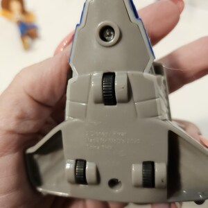 May include: A gray and blue toy spaceship with black wheels. The toy has a blue stripe along the top and is marked with text. The toy is a collectible item.